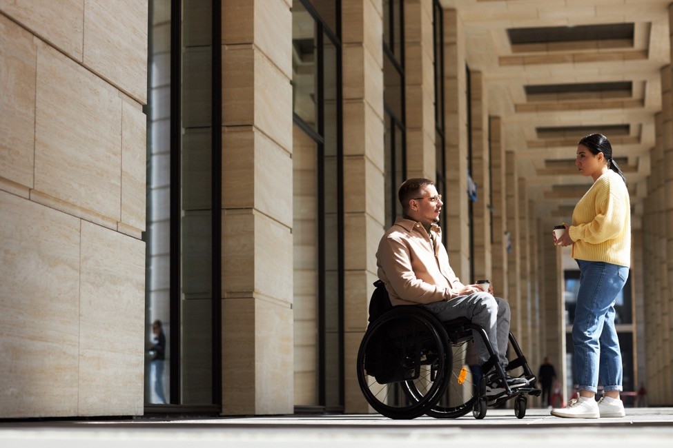 A man in a wheelchair talks to a woman outside a building. 