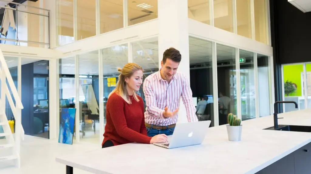 Two people looking at the laptop in a small office space with glass partitions and a staircase