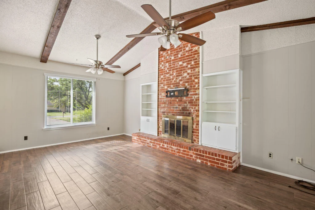 A room with a wooden floor, fan, shelves, fireplace, and a window