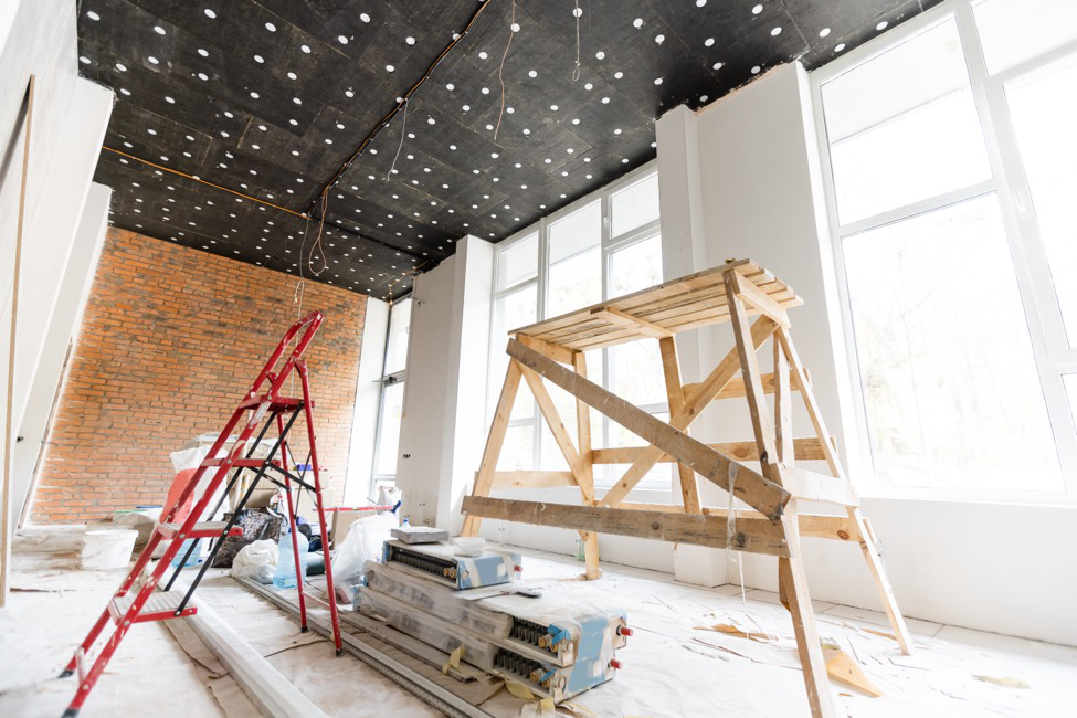 A tenant improvement project in process with a ladder, an elevated platform, a covered floor, white walls, and exposed brick